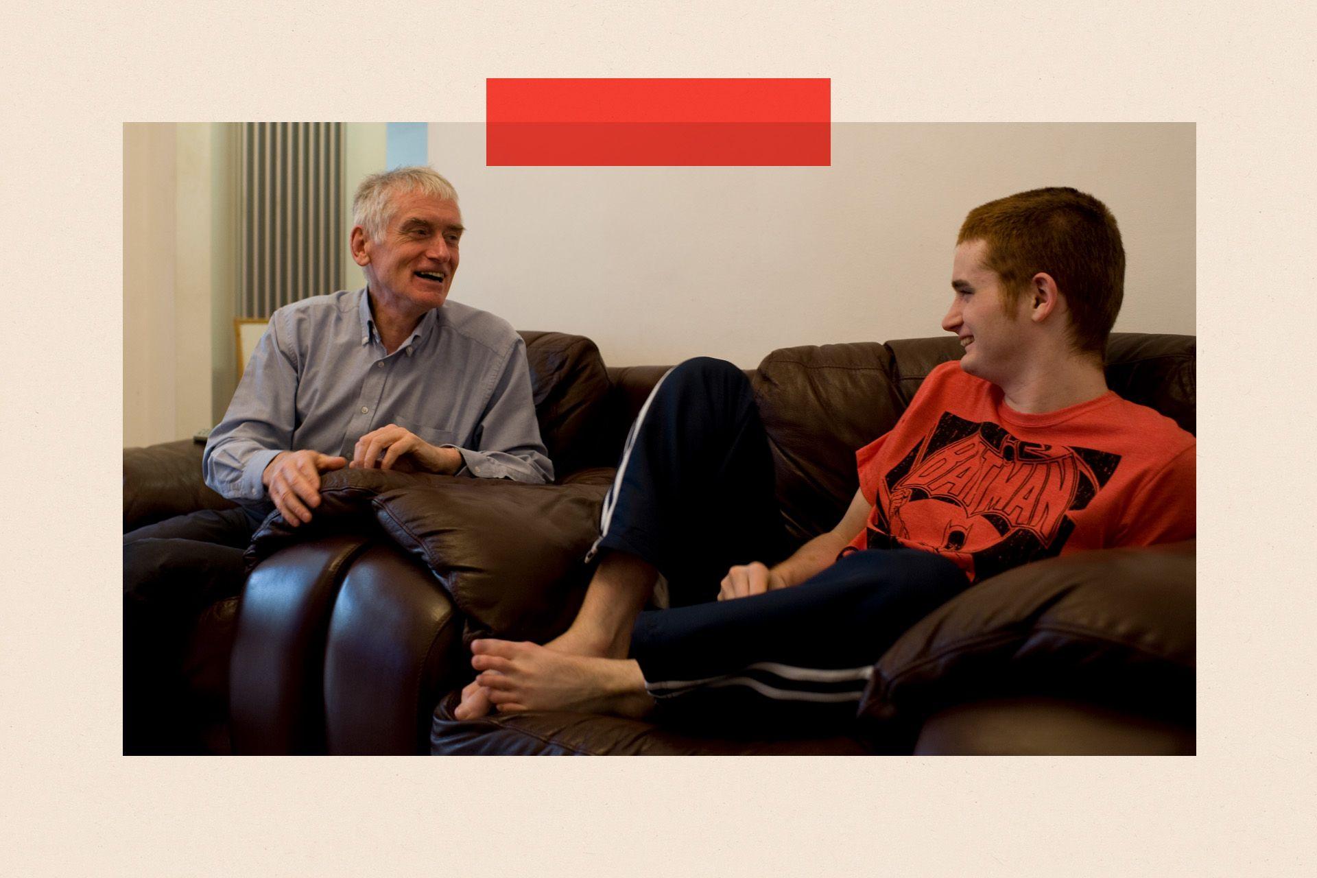 Michael and James Fitzpatrick sit on a sofa together. Michael wears a grey shirt and dark trousers, and james wears a red Batman t-shirt and dark tracksuit bottoms