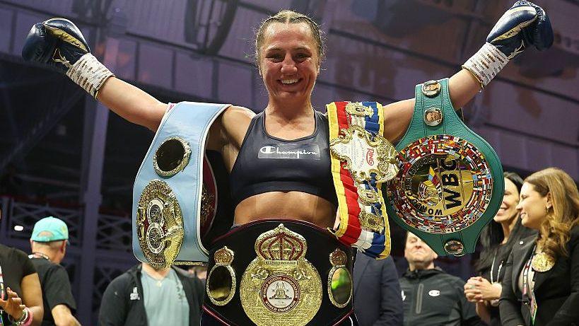 Ellie Scotney smiles and stretches her arms wide as she shows off the WBO, WBA, IBF and WBC super-bantamweight belts