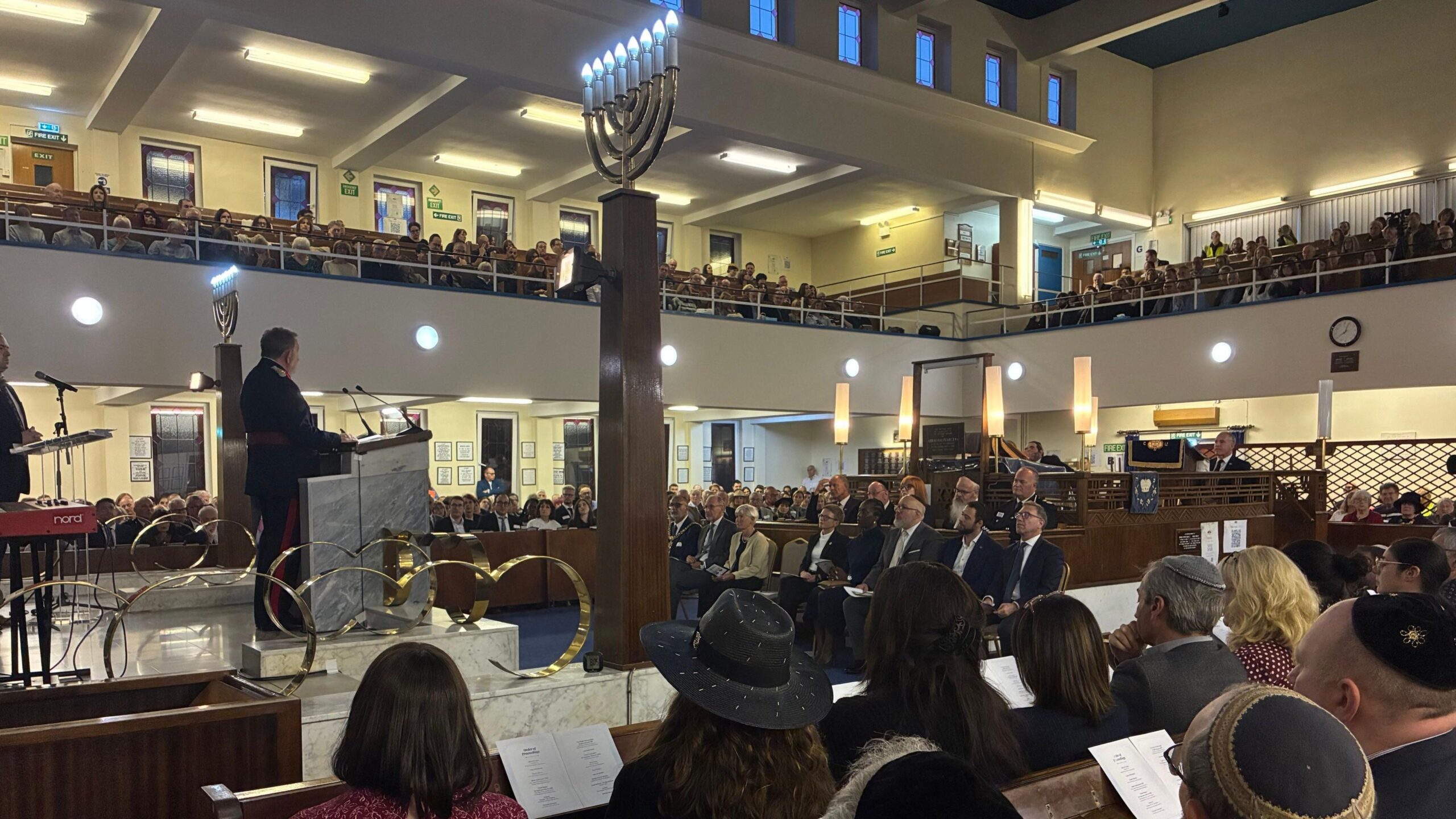 A man in a uniform is addressing the congregation while standing on a raised platform area, with two Menorahs on wooden pillars to either side of him. Many people can be seen in the seats of the shul.