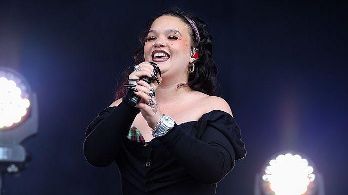 Singer Lola Young is pictured on stage at the Reading music festival. She is smiling while holding a microphone. She is wearing a black top with a tartan trim.