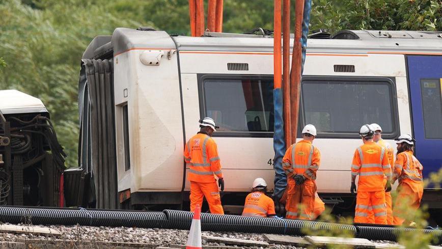 Recovery workers in orange high-visibility clothing and white safety helmets stand beside a Southern train carriage as lifting equipment is attached. Thick straps hang from a crane above the carriage, while cables, pipes and railway infrastructure are visible in the foreground.