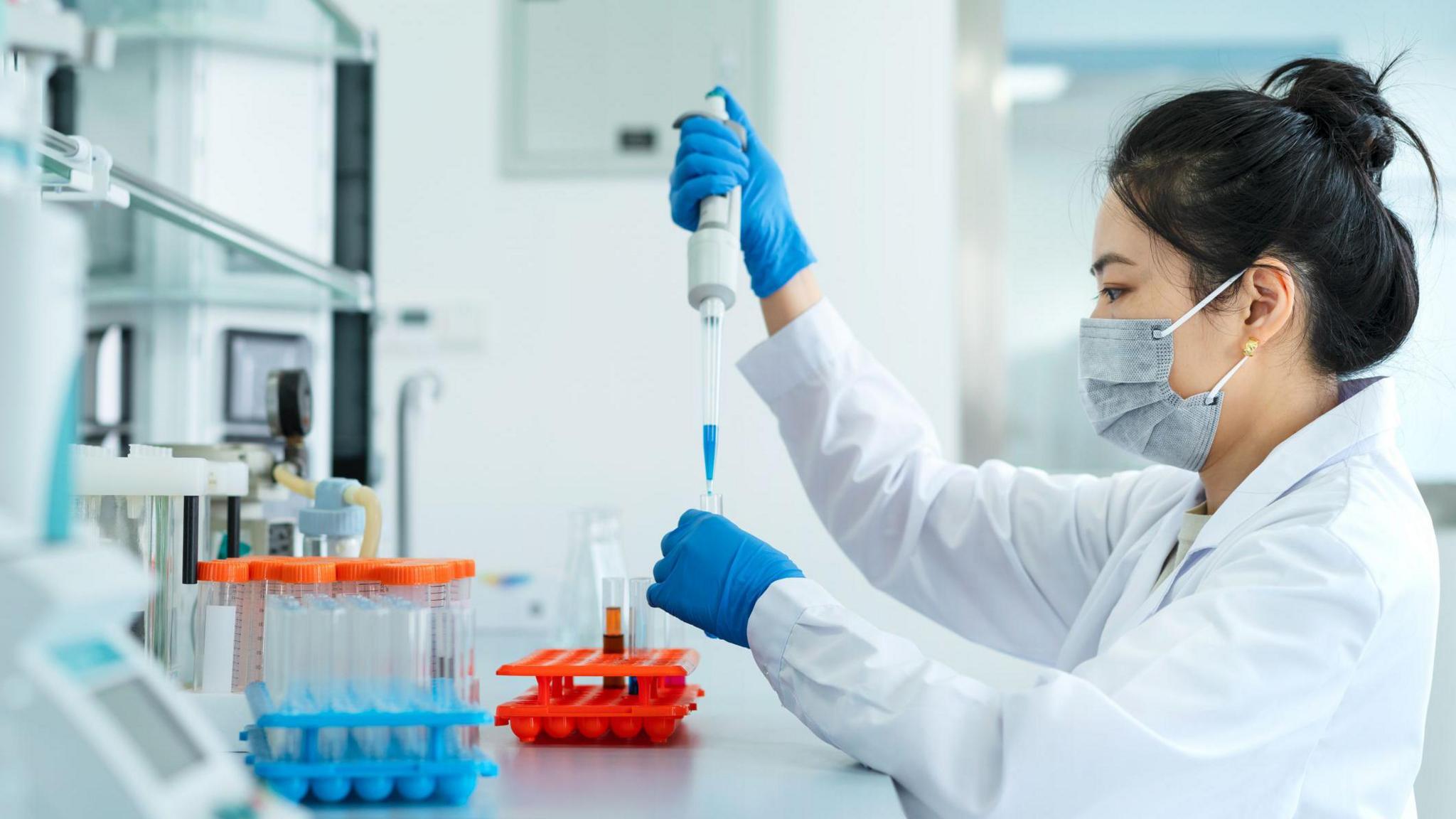 A laboratory setting with a person in a white lab coat working at a bench. The person is wearing protective gloves and a face mask while carefully using a pipette to transfer liquid into a small test tube. Several racks of test tubes with coloured liquids are arranged neatly on the work surface in the foreground. Scientific equipment and shelves are visible in the background, suggesting a clean and organised workspace