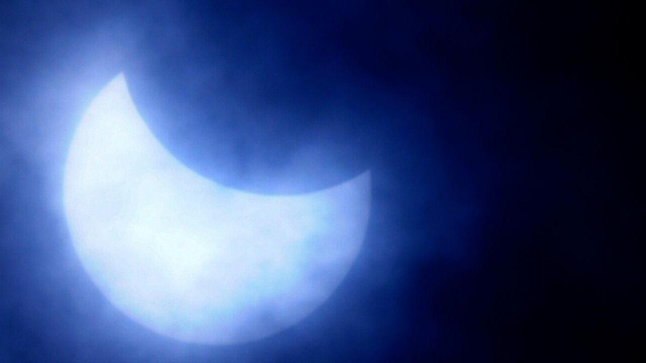 A view of the sun during a previous partial eclipse, which appears as a silvery-blue crescent against a black sky surrounded by a halo of light.