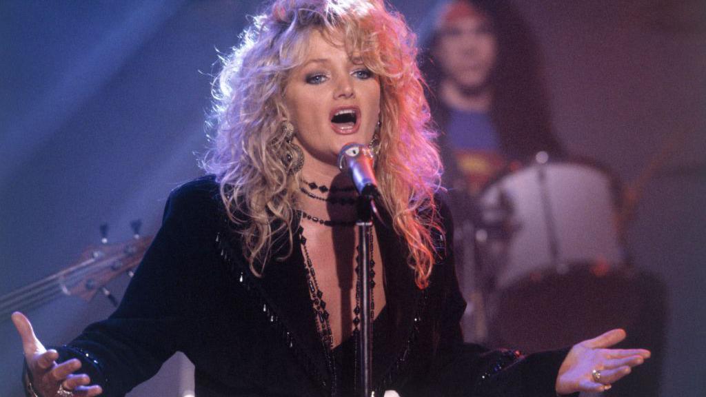 Bonnie Tyler performing on stage - she has blonde wavy hair and is wearing all black clothing. Her arms are outstretched and her mouth is open as she sings into a microphone