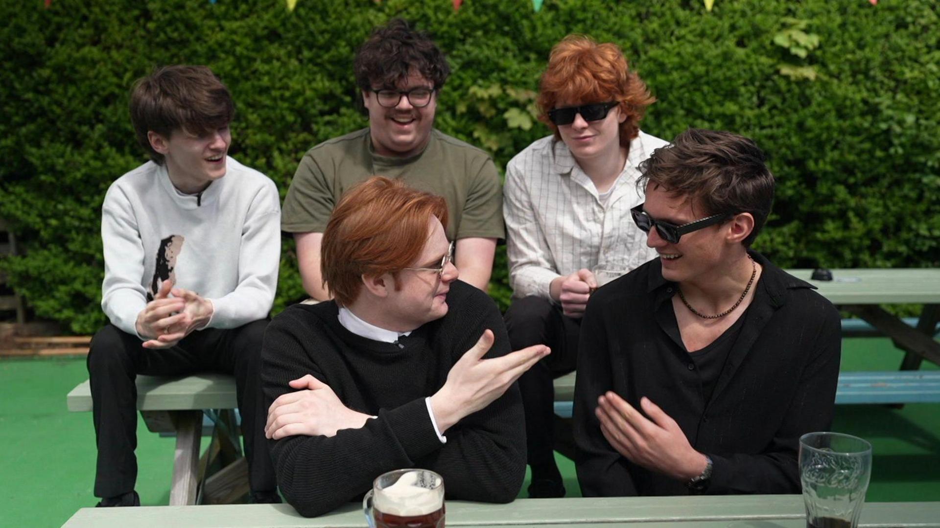 Swindled sit in the beer garden of the Board Inn pub in Sunderland. Felix is dressed in a white jumper, Thomas is wearing a green t-shirt, Will is wearing a white overshirt, Jonny is wearing a black jumper with a white collar and Alex is wearing a black shirt with a necklace.