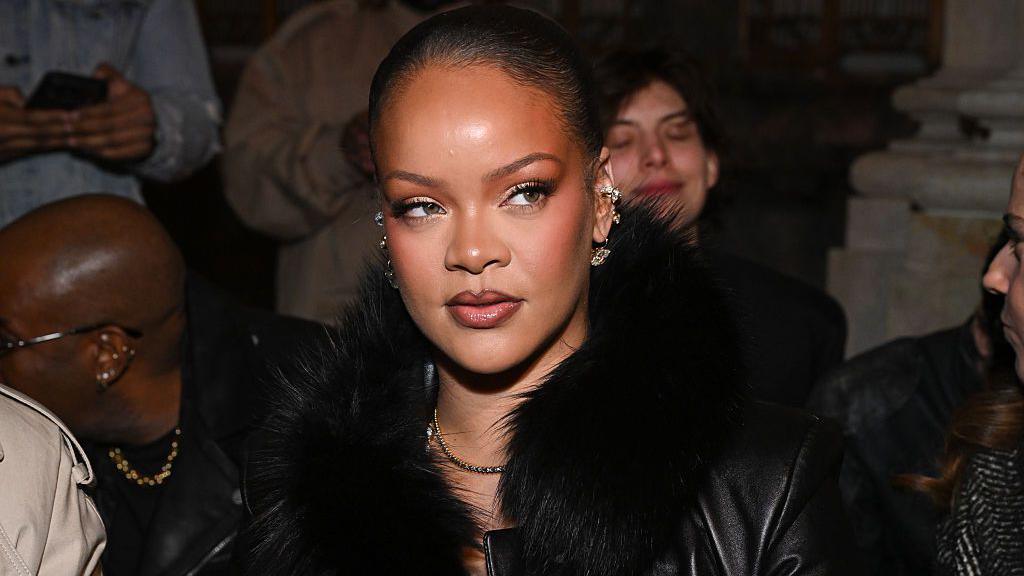 Rihanna looks to the side with her hair slicked back. She wears several earrings and a black fur collared jacket at the AWGE fashion show as part of RTW Fall 2026 on 13 February 2026 in New York, New York.