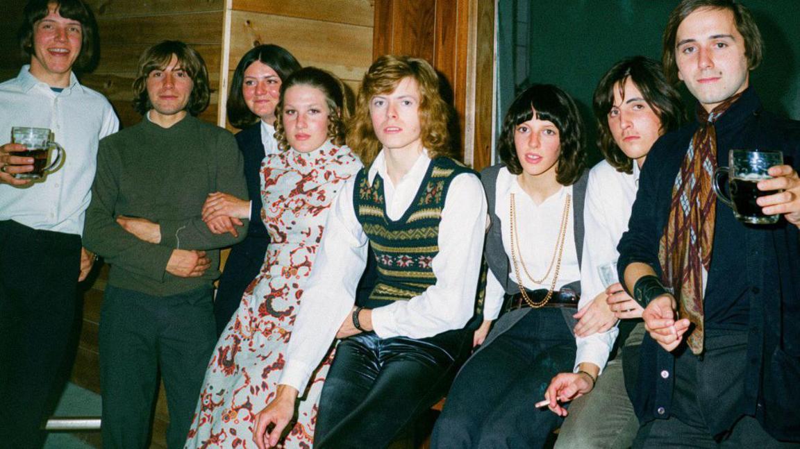 David Bowie, who has curly dark blonde hair, poses for a photo backstage at a club surrounded by seven teenagers including four boys and three girls.