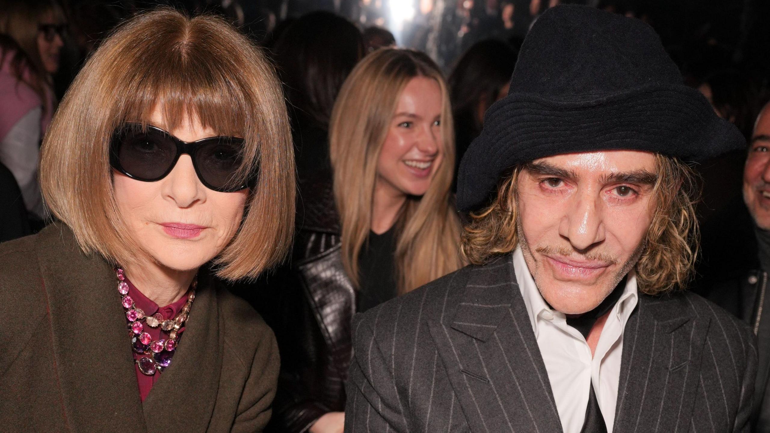 Anna Wintour in dark glasses and John Galliano in grey suit and black hat, seated together at a fashion show and looking expressionless at the camera