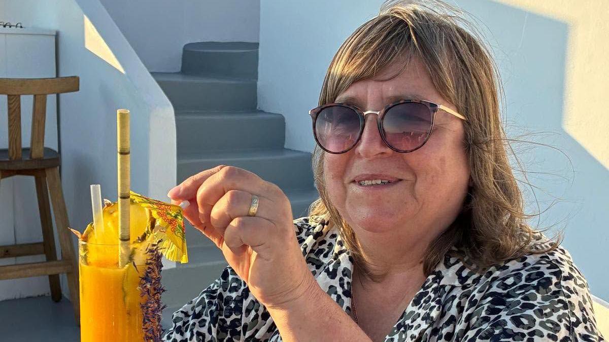 A woman wearing sunglasses and a leopard print shirt holding a yellow cocktail. She is smiling and looking into the camera.