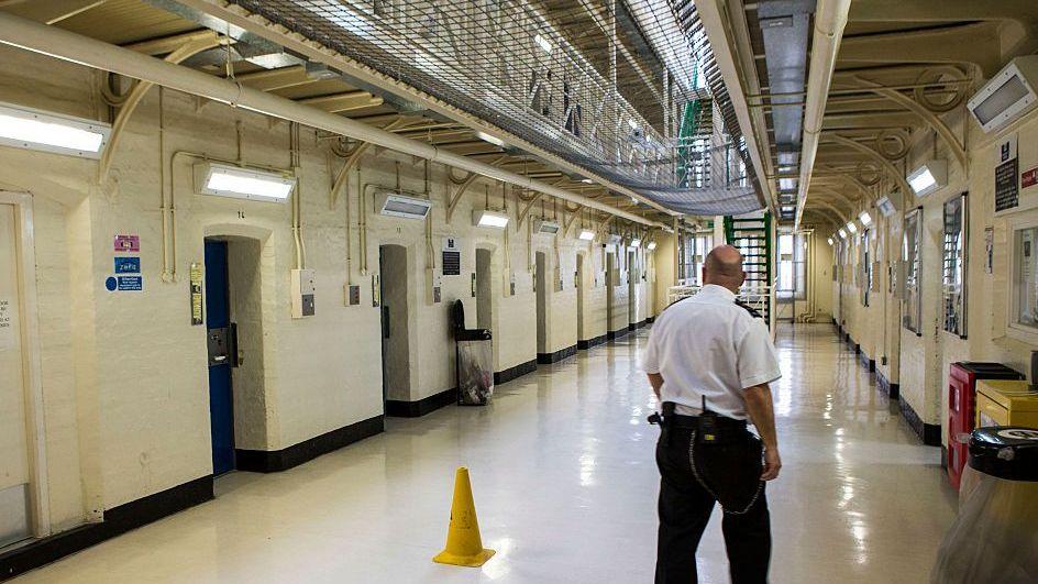 A prison guard in a white top and black trousers walks down a prison hallway. The walls and floor are white.