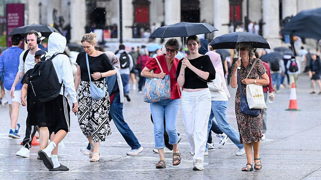 People hold umbrellas as they walk across Trafalgar Square during a heavy rain shower on August 20, 2026