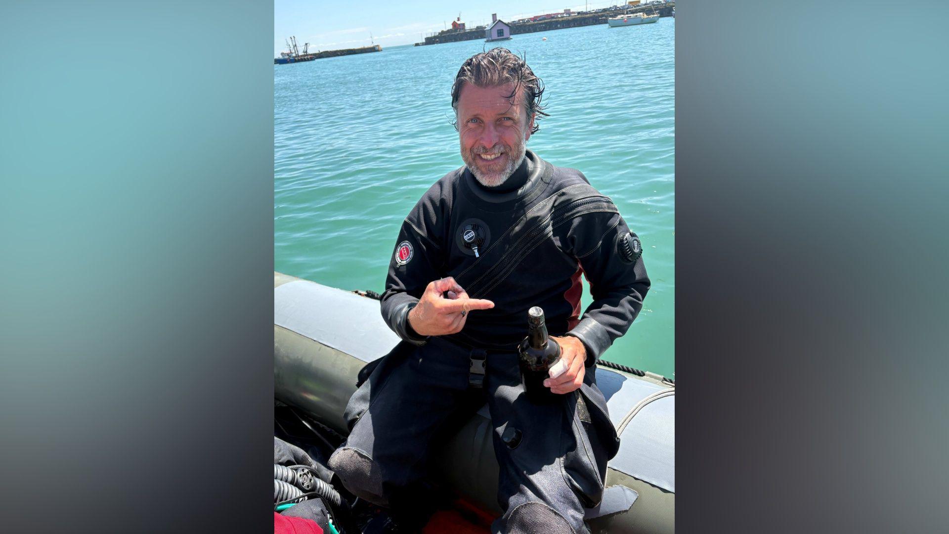 A man with dark hair, swept back, holding a black bottle. He is wearing a wet-suit and sitting on the side of a boat. He has a grey beard and is smiling at the camera, pointing at the bottle.