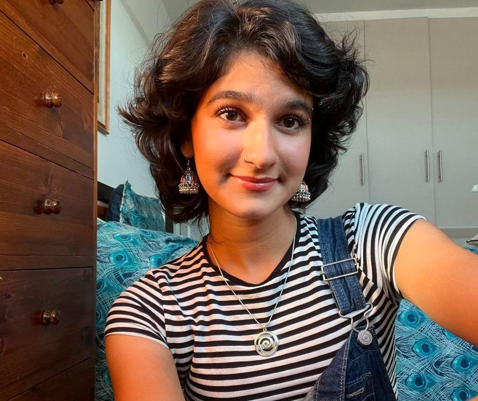 A woman in a striped black and white T-shirt sits in a bedroom and smiles at the camera