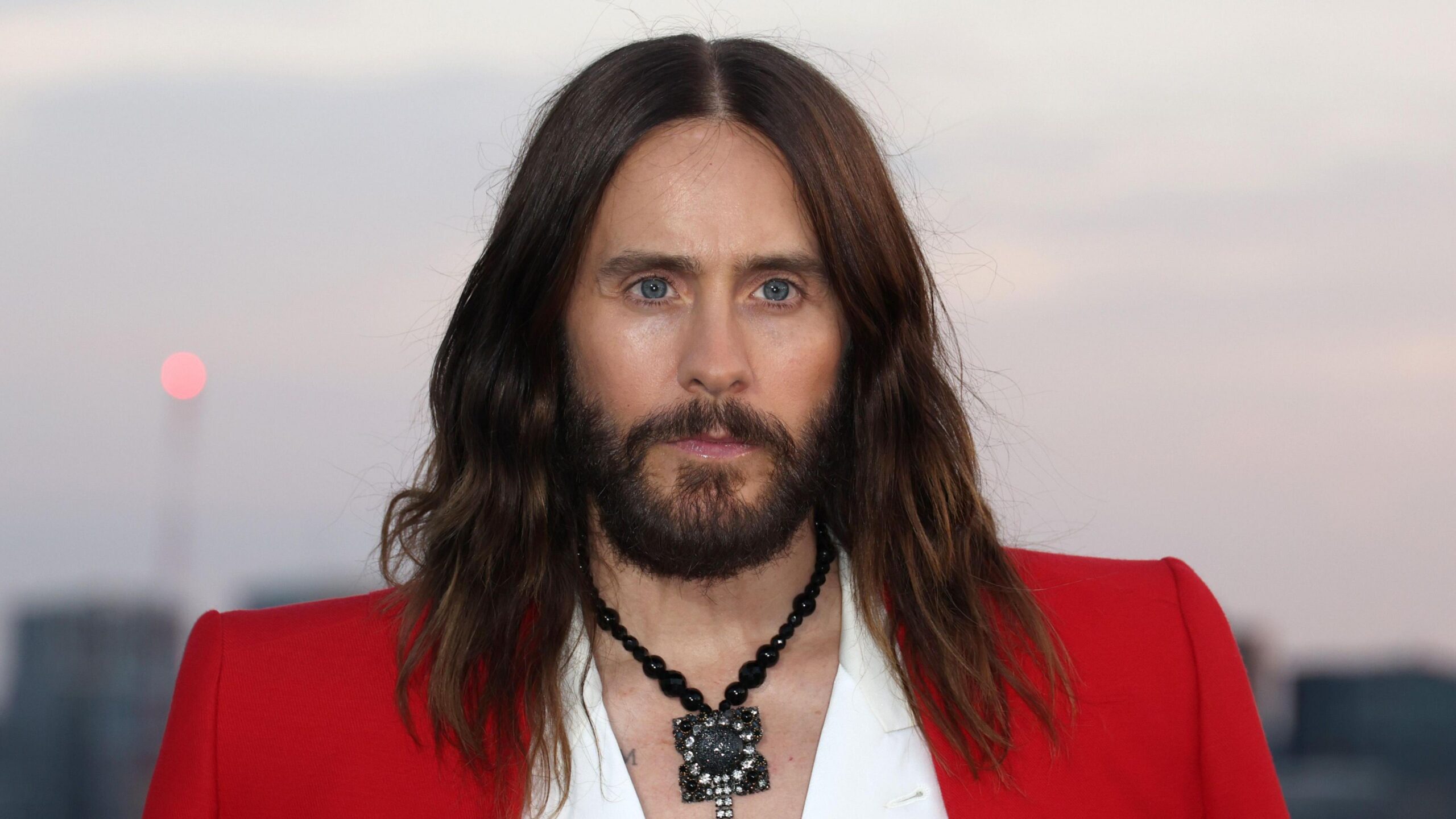 Jared Leto wears a red jacket over a white shirt. There is a large black necklace around his neck.