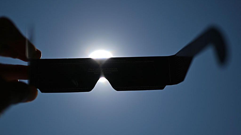 Hand holds up eclipse-viewing glasses up to a blue sky