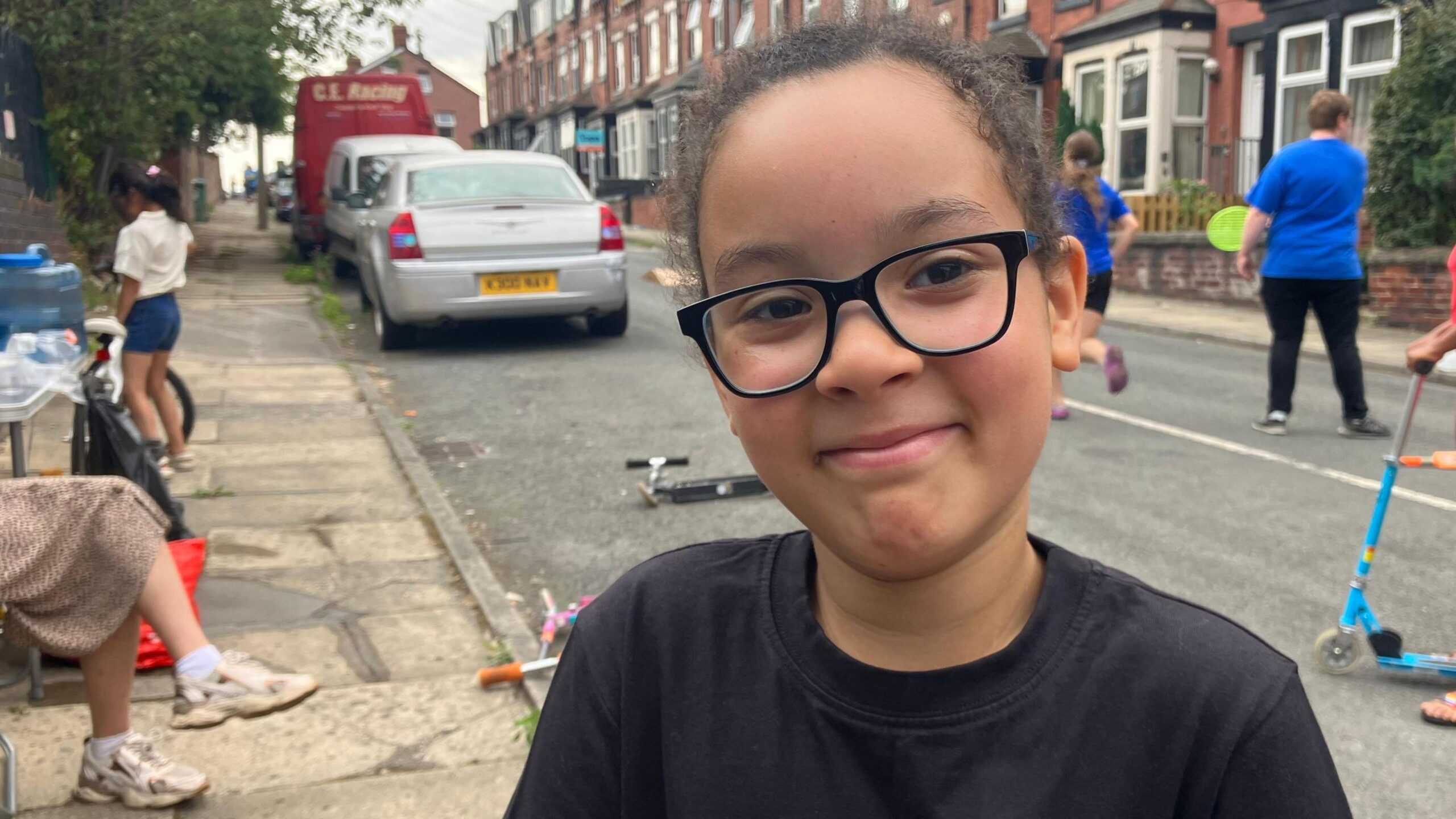A seven year old boy wearing a black t shirt and black glasses is smiling at the camera in the centre of frame. Behind him are children playing on scooters and with tennis bats and balls in a residential street, while parents sit on the pavement.