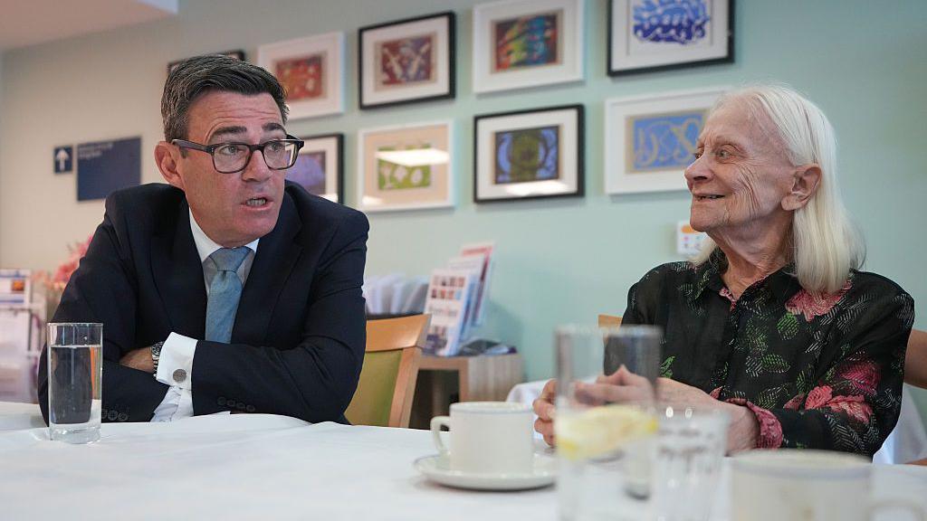 Andy Burnham sits at a table in a care home, talking face-to-face with a resident. The prime minister is in a dark suit and glasses and leans forward while speaking, and the other, with long white hair, sits opposite. Several glasses of water and cups are on the table, with framed artwork displayed on the wall behind them.