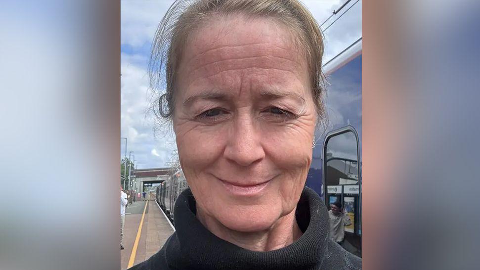 Sarah Nelson selfie on a railway station platform after having to come off a train. She has light brown hair worn back in a ponytail and wears a black polo neck jumper. She is smiling.