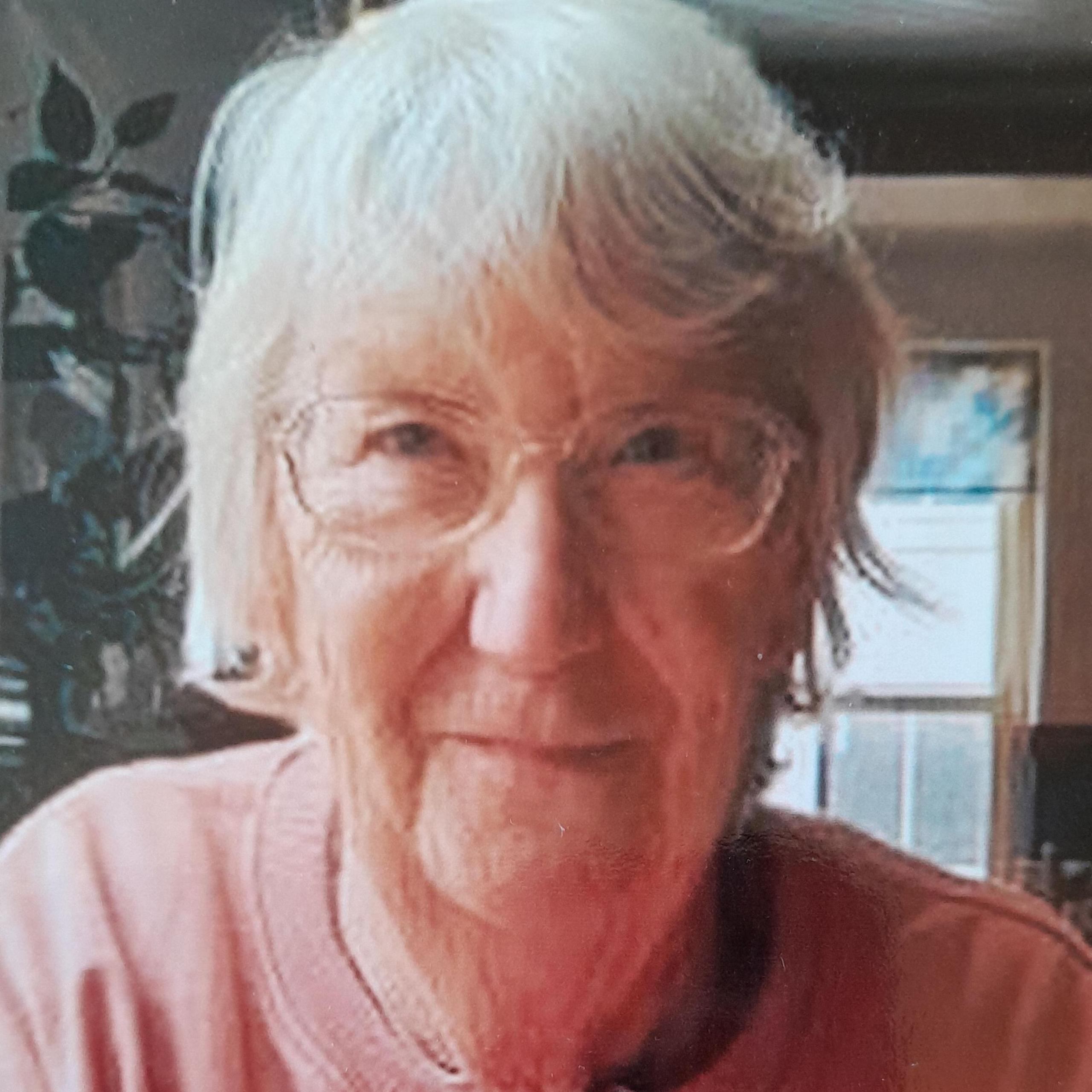 An older woman with short white hair and clear rimmed glasses looks into the camera . She wears a pale pink jumper with the words 'be yourself' on.