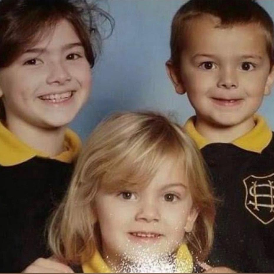 A school photo of three children in uniform. They are wearing dark jumpers over yellow polo shirts.