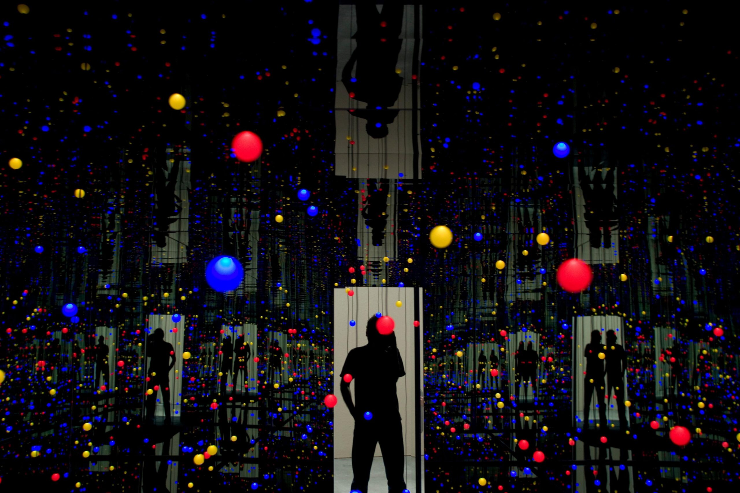 A Kusama installation - a dark space with many small coloured dots in the air, and reflections of light doorways with silhouetted figures
