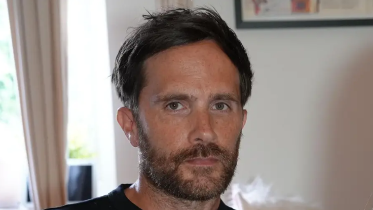 Craig Bowe sitting in a living room. He has dark hair and a brown beard. He looks serious in his expression while wearing a black T-shirt.