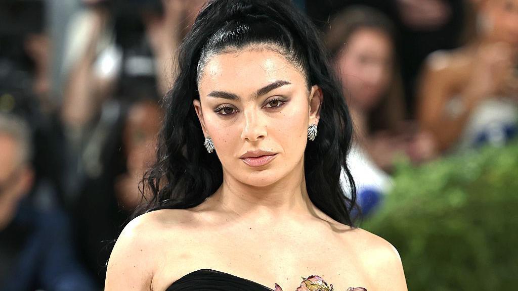 Charli XCX wears a black dress, with a flower on it. She has black hair, dark eyebrows and is looking directly at the camera lens.