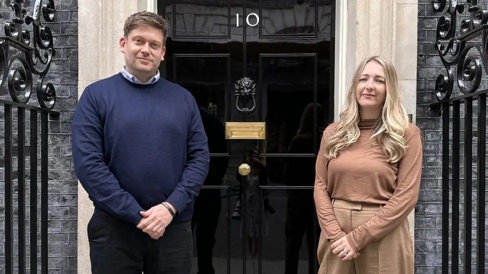 Frances and Ceri Menai-Davis standing side by side on a pavement directly in front of the black front door of a formal building. The door has a polished finish and features a brass letter slot and a lion-head door knocker at the centre. Above the door, the number “10” is clearly visible, indicating the entrance to 10 Downing Street in London. The doorway is framed by light stone columns,