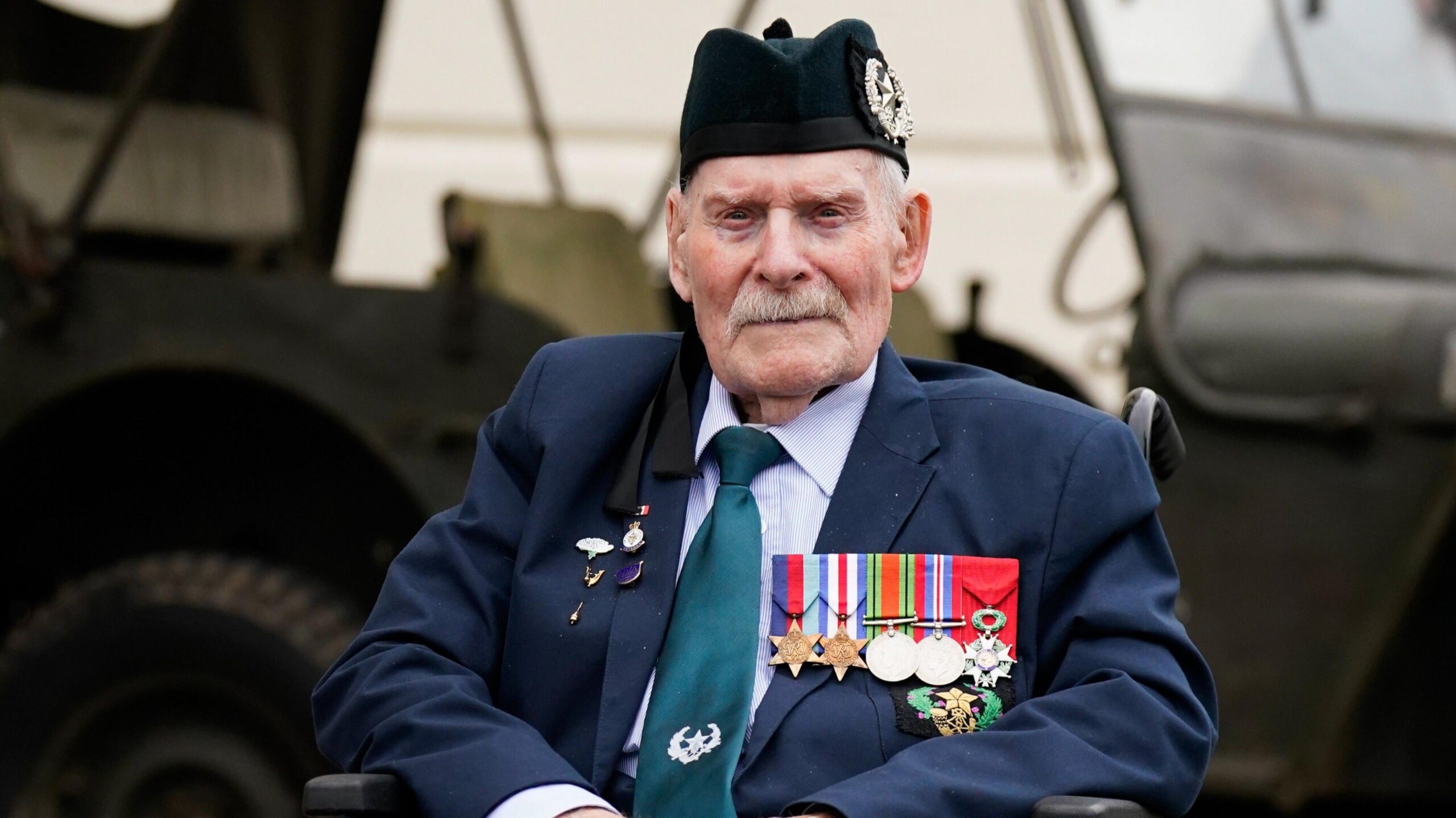 Donald Turrell is sitting in a wheelchair in front of a military vehicle. He is wearing a black army hat and a suit. On the left side of his jacket is a row of medals from World War Two.