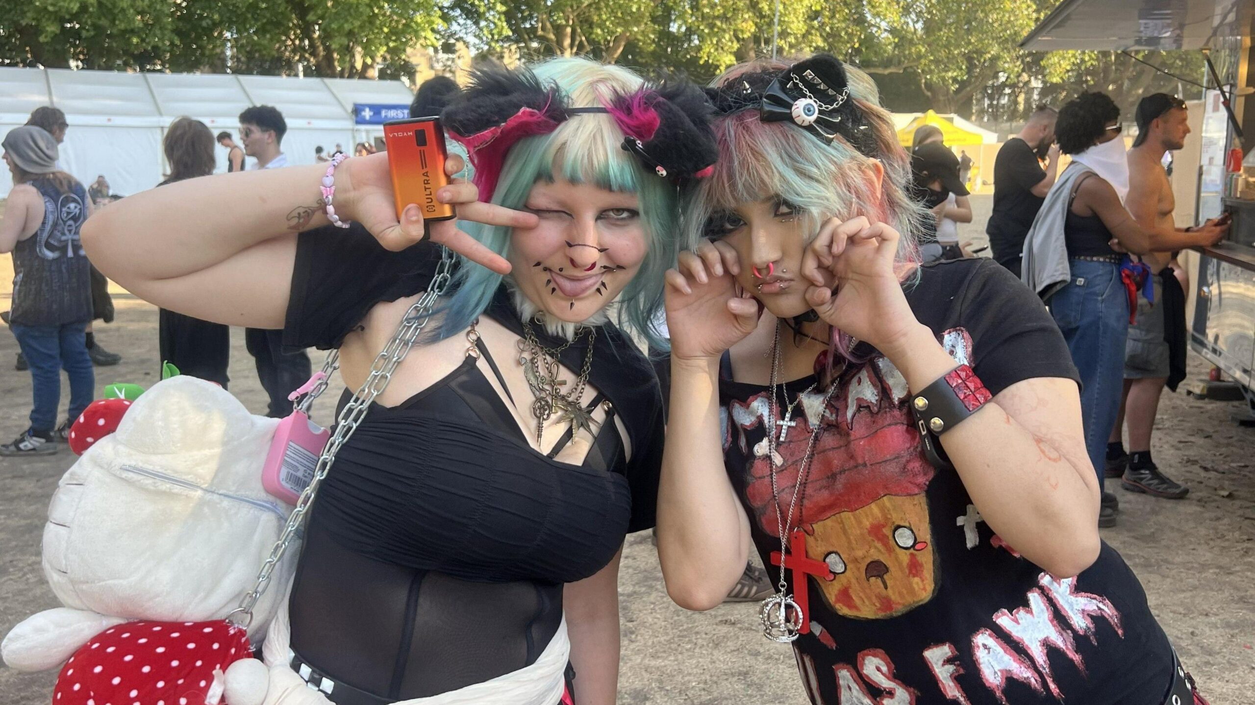 All Points East festival goers Bin and Zari smile at the camera wearing their festival gear. Bin wears a sheer black top and has green and blonde hair with a black furry headband. Zari wears a black slogan t-shirt and has pink and green hair.