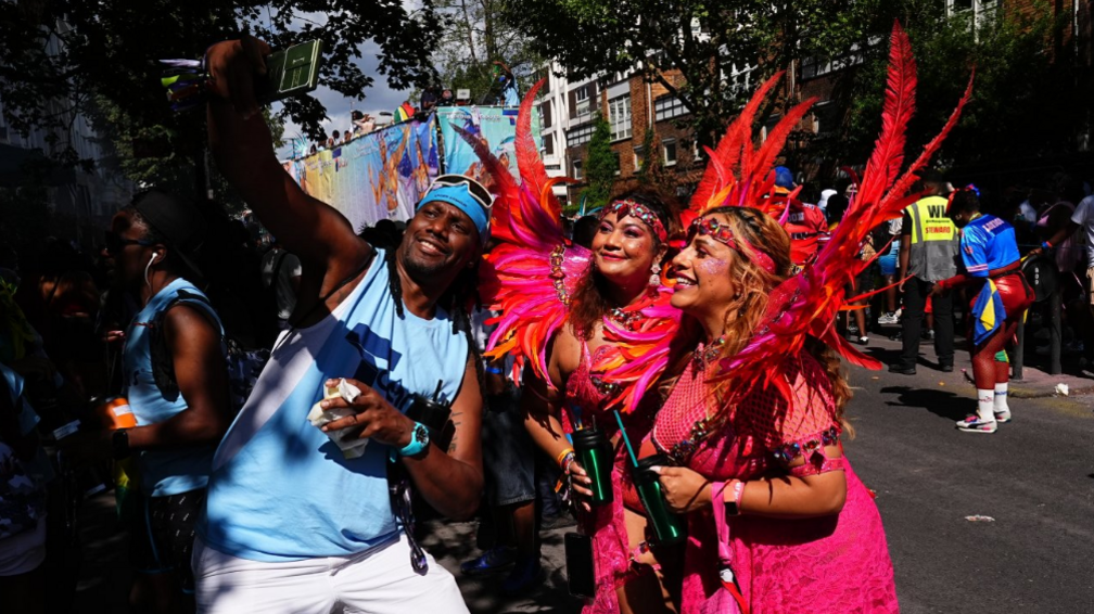 Two women in matching pink and orange costumes smile for a selfie image with a man in a blue vest top and white shorts.