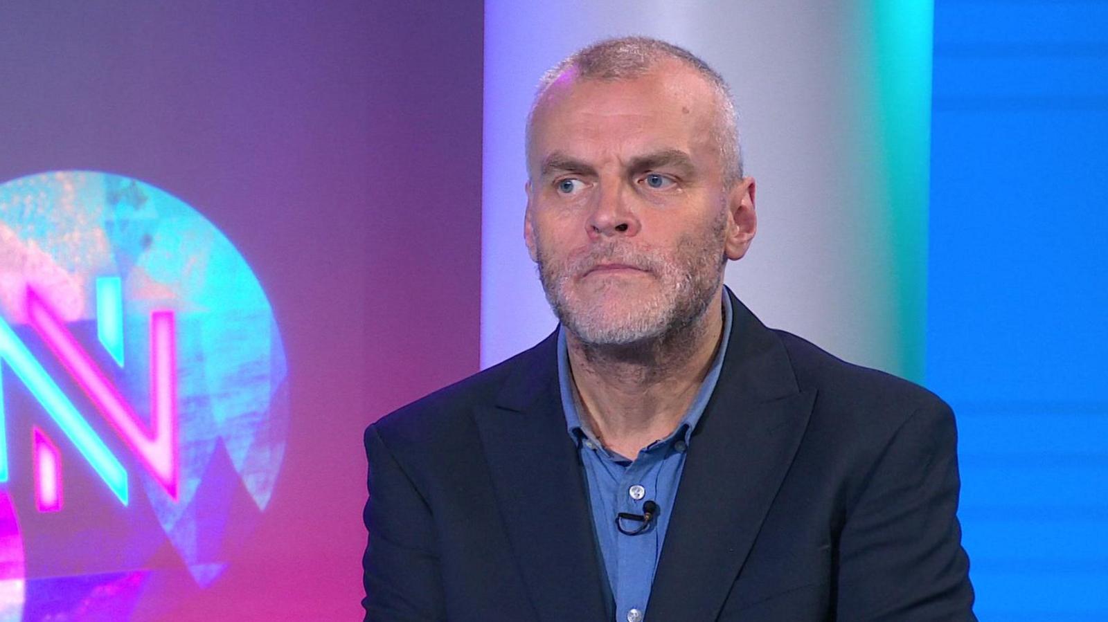 Tim Montgomerie, with short cropped hair and a grey beard, is wearing an open necked shirt and suit jacket. He is in the Newsnight studio