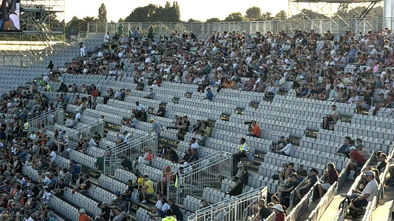 A photo of stands at a stadium. There are empty seats which are grey. You can also see fans sitting in different rows.