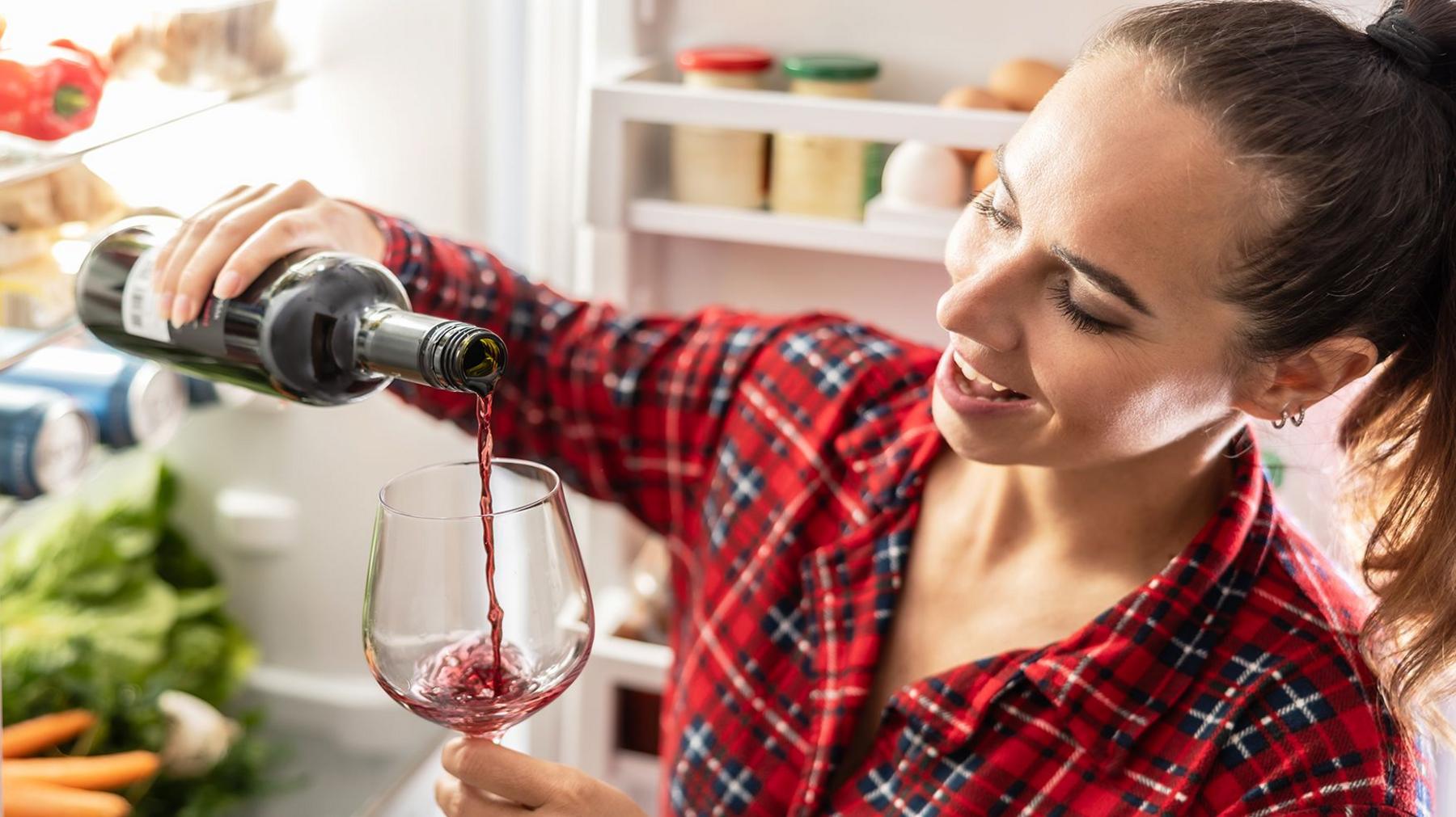 Wine bottle tilted over a wine glass as red wine is poured into the glass in the foreground. A person wearing a red checked shirt holds the bottle and glass. An open refrigerator is visible in the background, with shelves containing eggs, containers and vegetables.
