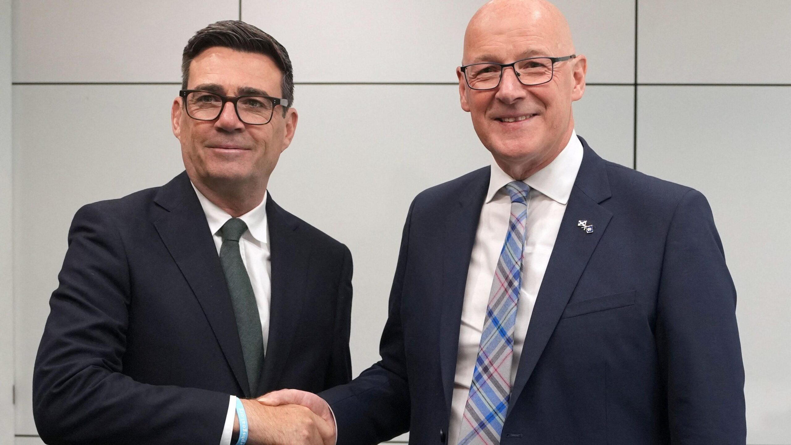 Andy Burnham, a man with thick dark hair and black-rimmed spectacles, is shaking hands with John Swinney, a man with a bald head and dark-rimmed spectacles. Burnham is in a dark blue suit with white shirt and solid green tie while Swinney is in a dark blue suit with white shirt and a blue, red and grey tartan tie. They are both smiling against a grey backdrop.