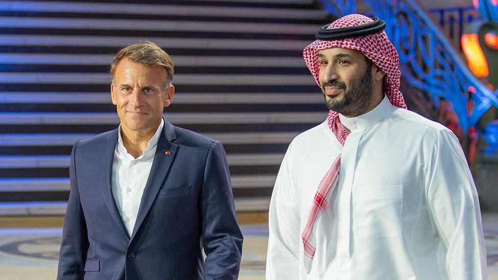 Emmanuel Macron and Prince Mohammed Bin Salman (MBS) stroll side-by-side into the venue of the EWC closing ceremony. Macron wears a navy blue suit, over a white shirt, collar open and no tie. MBS wears a traditional thawb (long, white robe) and red and white head-dress.