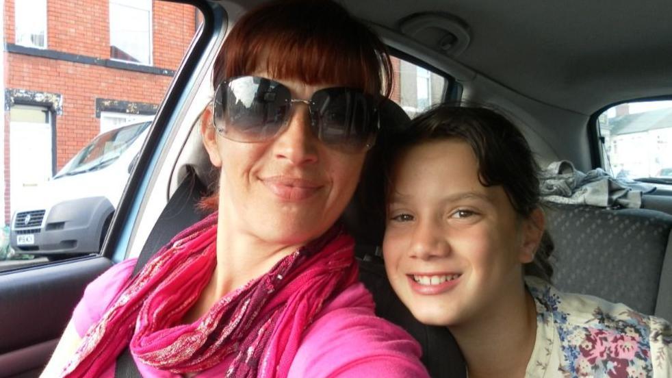 A woman with tied back dark hair and large sunglasses smiles at the camera in a pink scarf and pink cardigan. A little girl grins and leans lovingly on her shoulder.