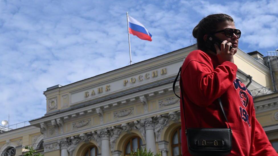 A woman wearing a red jumper and sunglasses, as well as a little bag on her right shoulder, talks on the phone in front of an exterior view of the Central Bank of the Russian Federation building on top of which the Russian flag can be seen