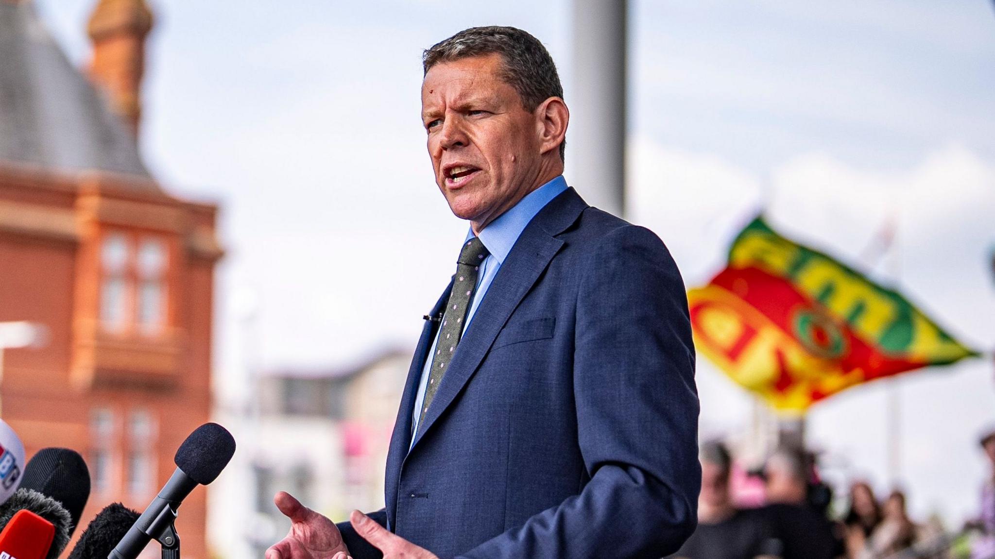 Rhun ap Iorwerth speaking into a set of microphones in front of the Pierhead building in Cardiff Bay, which is out of focus. A group of supporters can be seen behind him, also out of focus.