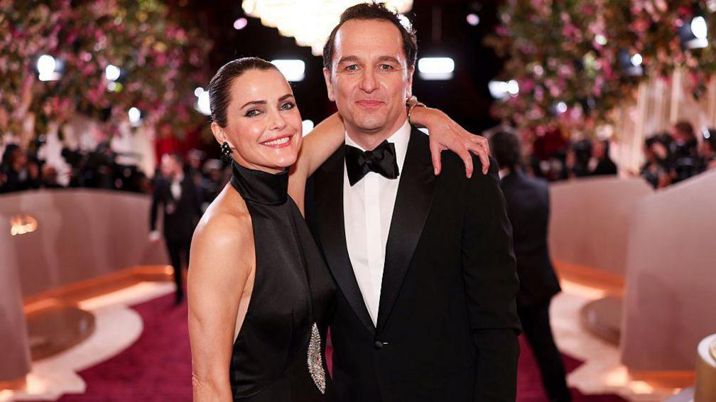 Rhys and Keri in a black tie formal wear on the red carpet, both smiling for the camera.