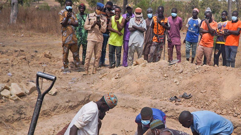 A line of men in masks watching grave diggers lowering bodies (not seen) into a grave in Kwara state. The top of a spade can be seen to the side.