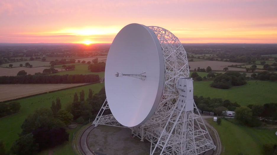 An aerial view at sunrise of the Lovell telescope at Jodrell Bank. Its huge white dish, taller than a house, is tilted upright on a steel cradle that sits on a circular track. Behind it, flat Cheshire farmland stretches to the horizon under a soft pink sky.