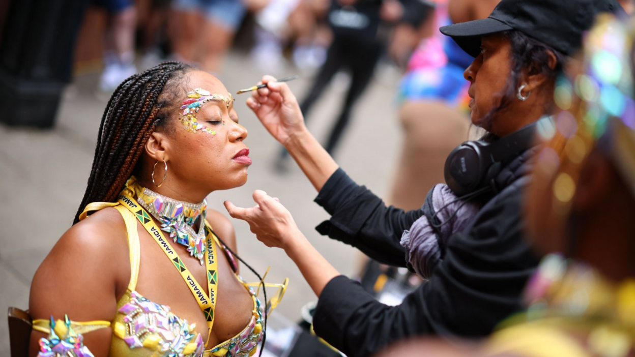 A woman in a yellow decorative outfit sits and has glitter and gems applied to her face.