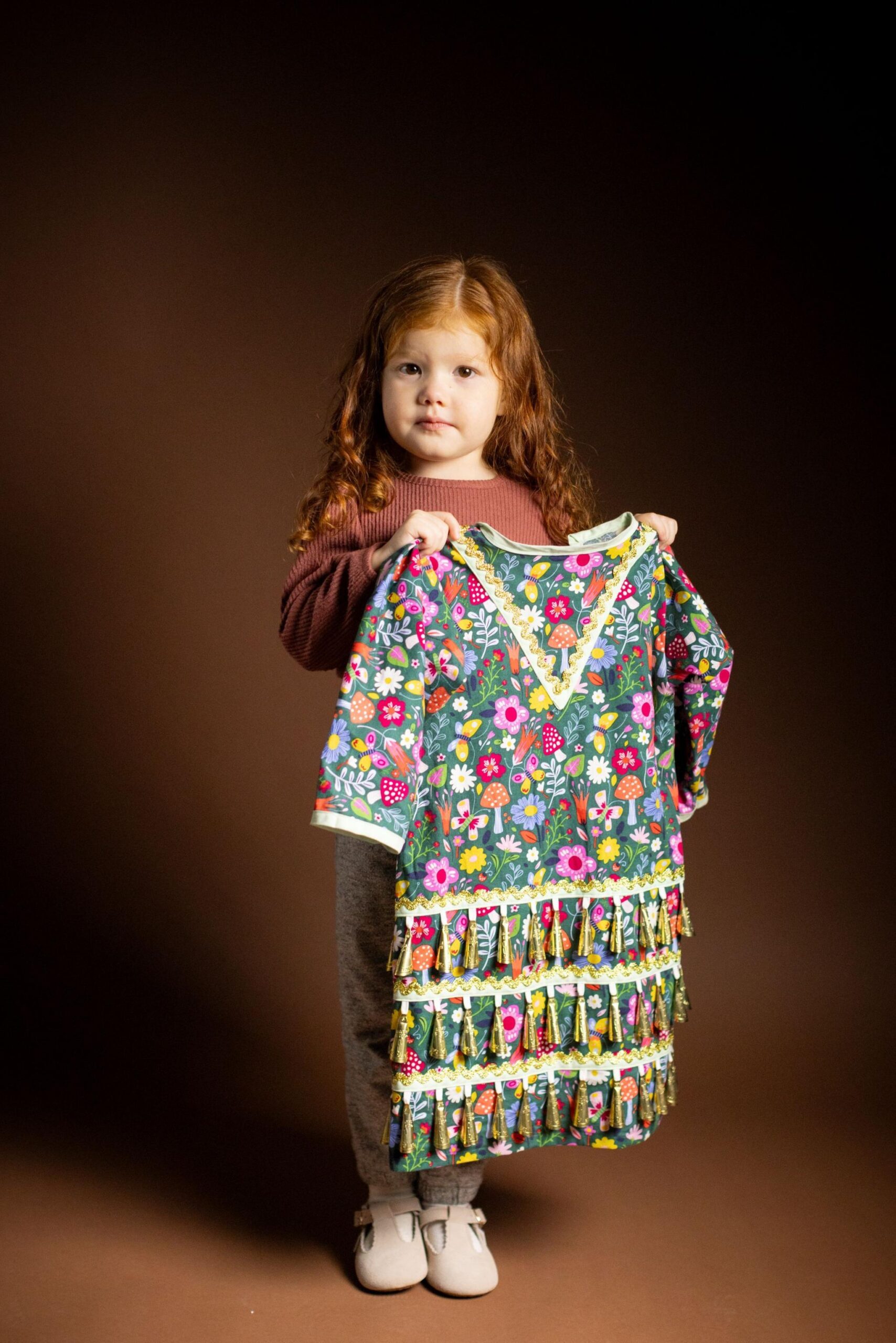 Full-length studio portrait of a young child with long curly hair standing against a dark brown background and holding up a colourful floral-patterned dress. The dress features bright flowers, mushrooms, and decorative tassel trim. The child wears a long-sleeved brown top, grey trousers, and light-coloured shoes. Warm lighting highlights the vibrant colours of the garment.