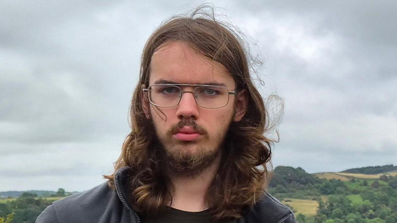 Tobias has long hair and glasses and is standing outdoors in front of rolling countryside under a cloudy, overcast sky.