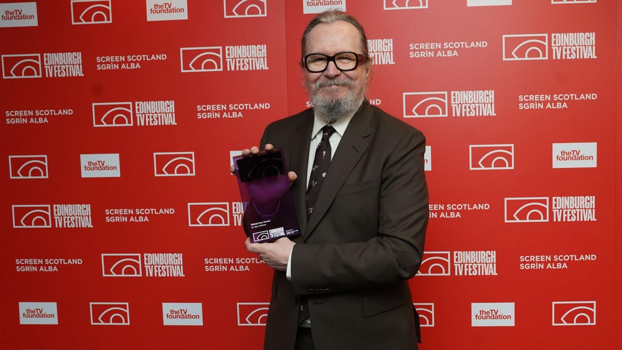 Sir Gary Oldman poses with his Global Icon award, a purple glass rectangle. Gary wears a chocolate brown suit, his hair is pulled back and he has a substantial beard in salt and pepper tones.