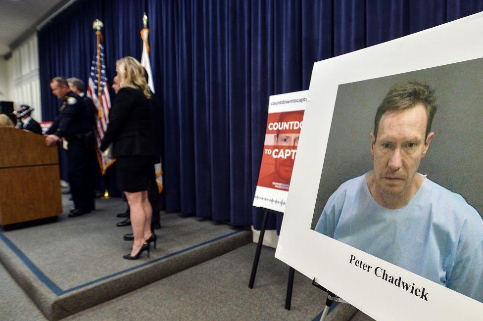 A row of police officers are on stage addressing a room of journalists (out of view) and there is a picture mounted on a board with the caption Peter Chadwick typed underneath his mugshot, where he's wearing a pale blue top.