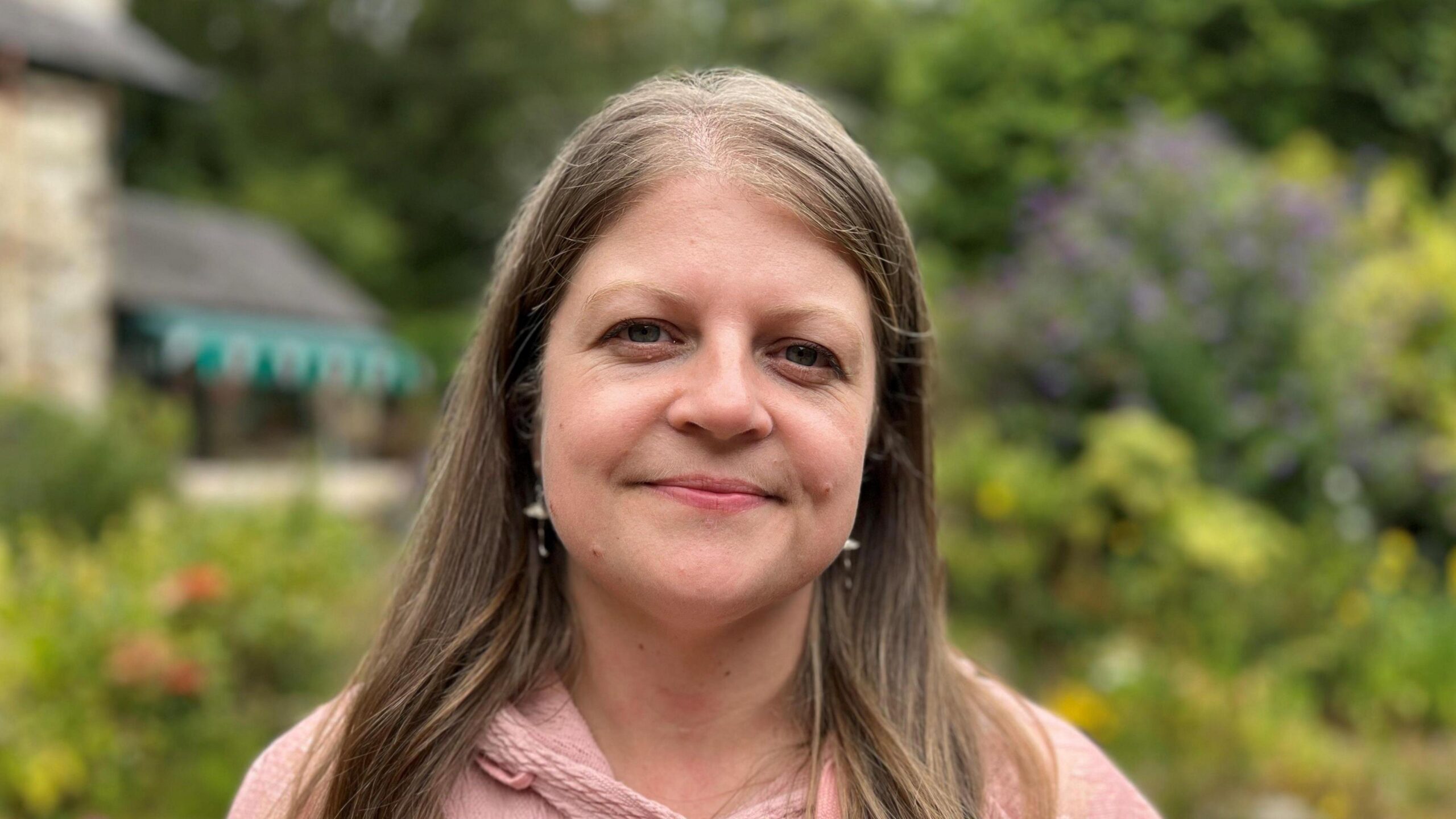 Evie Roberts looking at the camera. She is stood outside the Pensychnant Nature Reserve building and is wearing a pink hooded sweatshirt and has long dark hair.