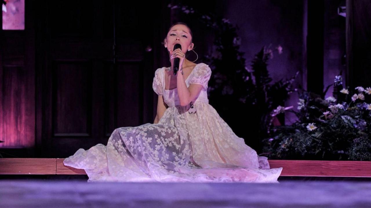 Ariana Grande sits on the stage, surrounded by flowers, as she sings on her Eternal Sunshine tour