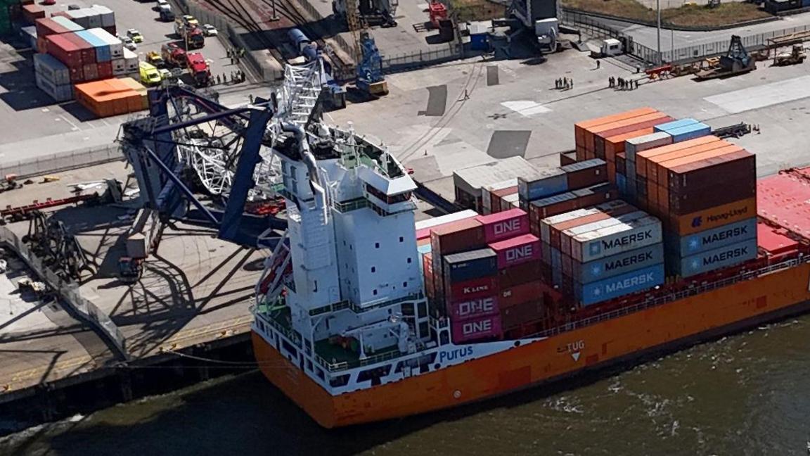 A crane has been knocked over on its side and is lay on top of an orange container ship. The crane is damaged, as are a number of containers on the vessel.